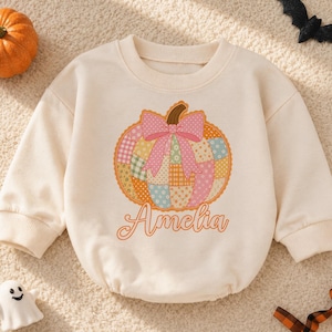 May include: A cream-colored long-sleeve sweatshirt featuring a central graphic of a patchwork-style pumpkin with a pink polka-dot bow. The name Amelia is printed in orange cursive script below the pumpkin design on the front of the garment.