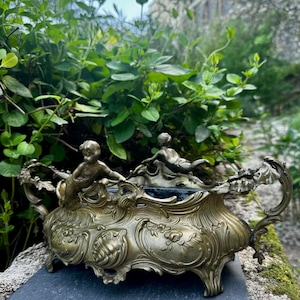 Może przedstawiać: Antyczny, brązowy pojemnik dekoracyjny z bogatymi detalami kwiatowymi i cherubinami. Pojemnik ma prostokątny kształt z zakrzywionymi uchwytami i stoi na czterech małych nóżkach. Tło przedstawia bujne zielone liście i kamienny mur.