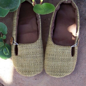 May include: A pair of beige woven fabric shoes with brown fabric lining and a button detail. The shoes are on a brown surface.