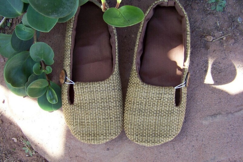 May include: A pair of beige woven fabric shoes with brown fabric lining and a button detail. The shoes are on a brown surface.