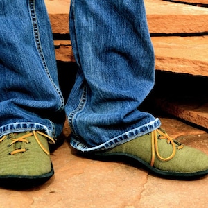 Puede incluir: Un par de zapatos de lona verde con cordones marrones y suela negra. Los zapatos son usados por una persona que lleva pantalones vaqueros azules.