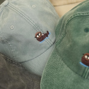 Könnte beinhalten: Eine Auswahl an Baseballkappen in verschiedenen Farben, darunter Grün, Blau und Beige. Jede Kappe hat ein gesticktes Otter-Design in Braun und Blau. Die Kappen sind aus einem weichen, strukturierten Stoff gefertigt.