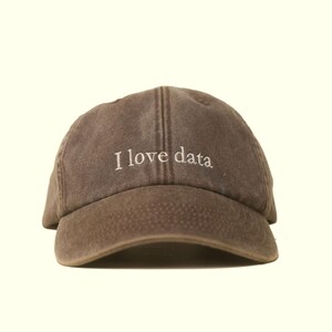 Puede incluir: Gorra de béisbol marrón con el texto bordado "I love data" en blanco. La gorra tiene una visera curva y una corona estructurada. El sombrero está hecho de un material suave y texturizado.