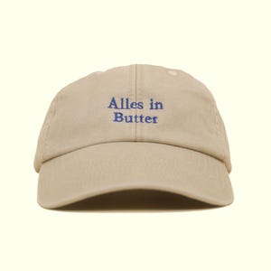 Op de afbeelding: Een beige baseballpet met de blauwe tekst "Alles in Butter". De pet heeft een gebogen klep en een klassiek ontwerp met zes panelen, geschikt voor dagelijks gebruik.