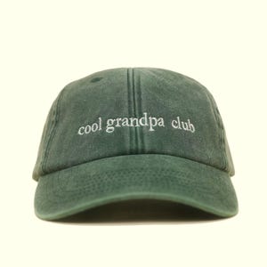 Könnte beinhalten: Dunkelgrüne Baseballkappe mit dem gestickten Text "cool grandpa club" in Weiß. Die Kappe hat einen gebogenen Schirm und ein klassisches sechsteiliges Design. Der Hut ist aus einem weichen, strukturierten Stoff gefertigt.