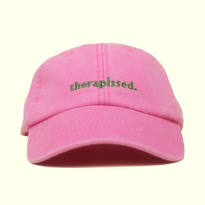 Könnte beinhalten: Eine pinkfarbene Baseballkappe mit dem grün gestickten Wort "therapissed.". Die Kappe hat einen gebogenen Schirm und ein klassisches Sechs-Panel-Design. Der Hut ist aus einem weichen, strukturierten Stoff gefertigt.