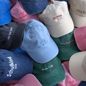 Könnte beinhalten: Eine Ansammlung von Baseballkappen in verschiedenen Farben, darunter Blau, Rosa und Grün. Mehrere Kappen haben gestickten Text, wie "EAT THE RICH", "raised by boomers" und "Holy Aperoly". Die Kappen sind aus einem weichen Stoff gefertigt.