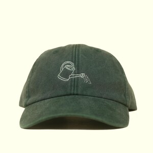 Könnte beinhalten: Eine dunkelwaldgrüne Baseballkappe mit einer weißen, gestickten Linienzeichnung einer Gießkanne auf der Vorderseite. Die Kappe hat eine strukturierte Krone, einen gebogenen Schirm und sichtbare Nähte entlang der Stoffbahnen.