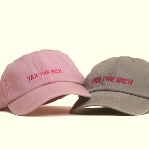 Könnte beinhalten: Eine hellrosa Baseballkappe und eine graue Baseballkappe, beide mit dem gestickten Text TAX THE RICH in rotem Garn. Jede Kappe hat eine strukturierte Krone, einen gebogenen Schirm und sichtbare Nähte über die Stoffbahnen hinweg.