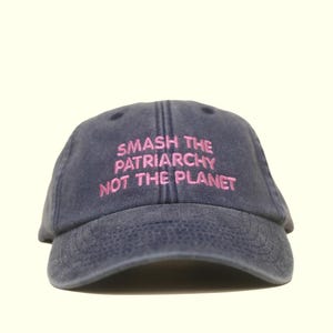 Könnte beinhalten: Marineblaue Baseballkappe mit dem pinkfarbenen, gestickten Text "SMASH THE PATRIARCHY NOT THE PLANET". Die Kappe hat einen gebogenen Schirm und einen verstellbaren Riemen. Der Hut ist aus einem weichen, strapazierfähigen Stoff gefertigt.