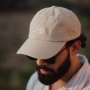 Könnte beinhalten: Beige Baseballkappe mit weißem, gesticktem Sonnendesign. Die Kappe hat einen gebogenen Schirm und eine klassische Sechs-Panel-Konstruktion. Der Hut ist aus einem weichen, strapazierfähigen Stoff gefertigt.