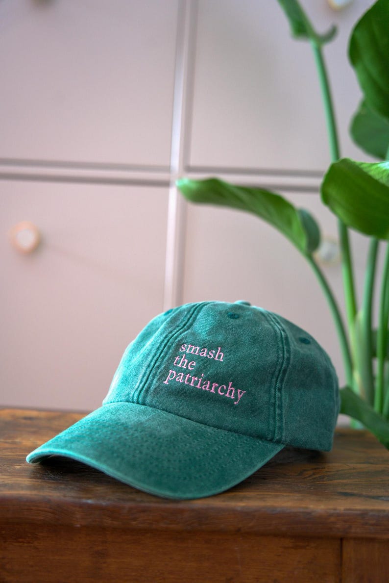 Op de afbeelding: Een tealkleurige baseballpet met de roze geborduurde tekst "smash the patriarchy". De pet is gemaakt van een zacht materiaal en heeft een gebogen klep. De pet ligt op een houten oppervlak.