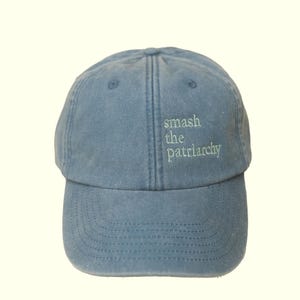 Op de afbeelding: Lichtblauwe baseballcap met de geborduurde tekst "smash the patriarchy" in groen. De cap heeft een gebogen klep en een gestructureerde kroon, geschikt voor casual wear.