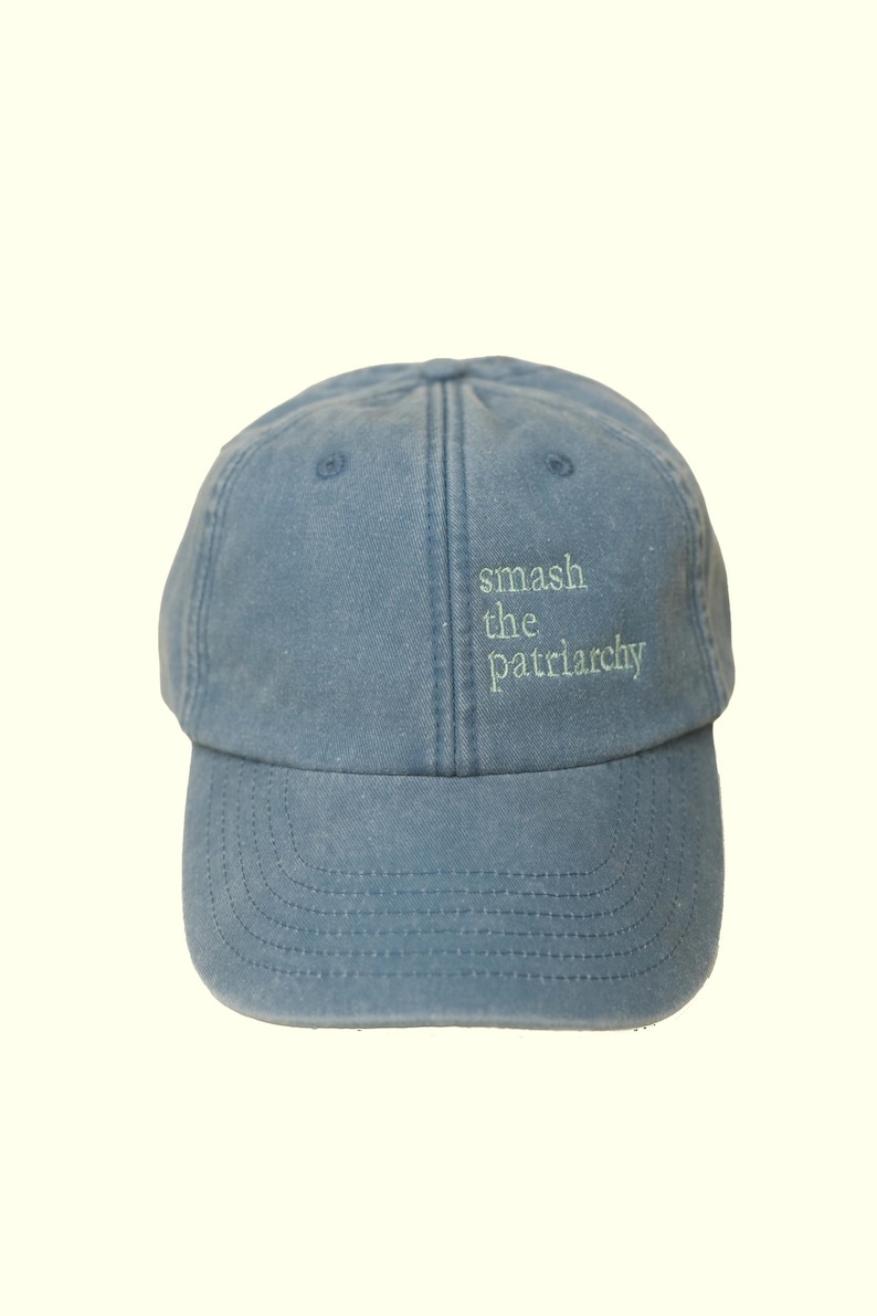 Op de afbeelding: Lichtblauwe baseballcap met de geborduurde tekst "smash the patriarchy" in groen. De cap heeft een gebogen klep en een gestructureerde kroon, geschikt voor casual wear.