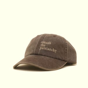 Op de afbeelding: Een bruine baseballcap met de geborduurde tekst "smash the patriarchy" in lichtbruine kleur. De cap heeft een gebogen klep en een klassiek zes-panelen ontwerp.