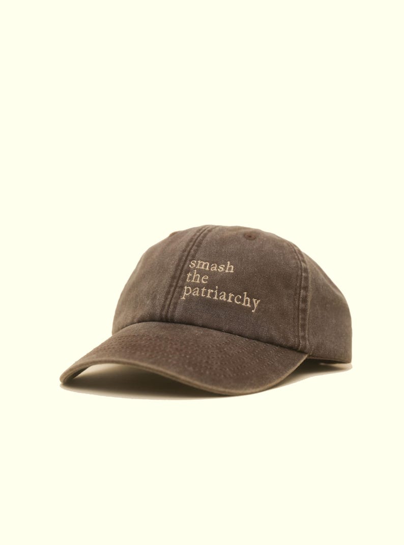 Op de afbeelding: Een bruine baseballcap met de geborduurde tekst "smash the patriarchy" in lichtbruine kleur. De cap heeft een gebogen klep en een klassiek zes-panelen ontwerp.