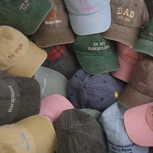 Könnte beinhalten: Eine Sammlung von Baseballkappen in verschiedenen Farben wie Hellblau, Rosa, Beige, Dunkelgrau und Grün. Jede Kappe ist mit einzigartigen gestickten Texten oder Grafiken versehen, darunter "LATE BUT CUTE", "DAD EST. 2025", "IN MY GARDEN ERA" und "therapissed".