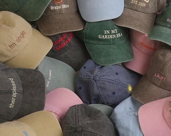 Könnte beinhalten: Eine Sammlung von Baseballkappen in verschiedenen Farben wie Hellblau, Rosa, Beige, Dunkelgrau und Grün. Jede Kappe ist mit einzigartigen gestickten Texten oder Grafiken versehen, darunter "LATE BUT CUTE", "DAD EST. 2025", "IN MY GARDEN ERA" und "therapissed".