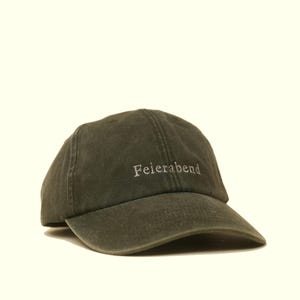 Könnte beinhalten: Dunkel olivgrüne Baseballkappe mit dem Wort "Feierabend" in einem helleren Grünton gestickt. Die Kappe hat einen gebogenen Schirm und ein klassisches Sechs-Panel-Design.