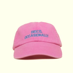 Könnte beinhalten: Eine pinkfarbene Baseballkappe mit dem gestickten Text "DISCO, OCCASIONALLY" in Blau. Die Kappe ist aus einem weichen Stoff gefertigt und hat einen gebogenen Schirm. Der Hut hat eine einfarbige rosa Farbe.