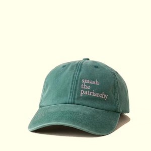 Op de afbeelding: Een teal baseball cap met de tekst "smash the patriarchy" in roze letters. De cap heeft een gebogen klep en een stoffen bandsluiting. De hoed is gemaakt van een zacht, versleten materiaal.