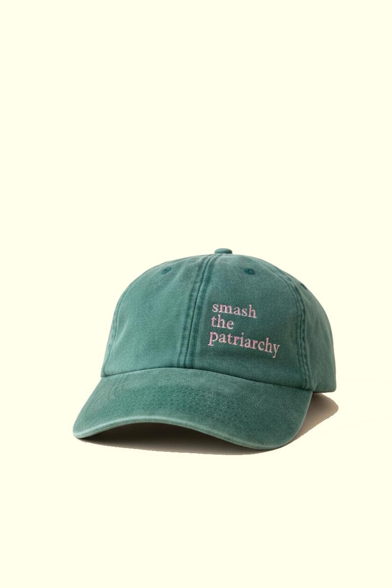 Op de afbeelding: Een teal baseball cap met de tekst "smash the patriarchy" in roze letters. De cap heeft een gebogen klep en een stoffen bandsluiting. De hoed is gemaakt van een zacht, versleten materiaal.