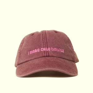 Könnte beinhalten: Weinrote Baseballkappe mit dem in Rosa gestickten Schriftzug "I make cute babies". Die Kappe hat einen gebogenen Schirm und ein sechsteiliges Design mit einem oberen Knopf und genähten Details.