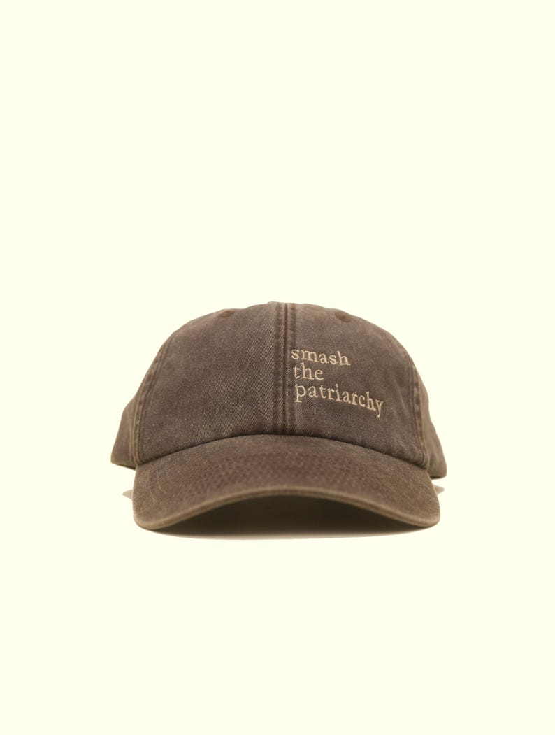 Op de afbeelding: Bruine baseballcap met de geborduurde tekst "smash the patriarchy" in een lichtere bruine kleur. De cap heeft een gebogen klep en een klassiek zes-panelen ontwerp.