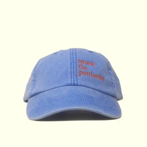 Op de afbeelding: Lichtblauwe baseballcap met de tekst "smash the patriarchy" in oranje geborduurd. De cap heeft een gebogen klep en een zes-panelen ontwerp, geschikt voor casual gebruik.