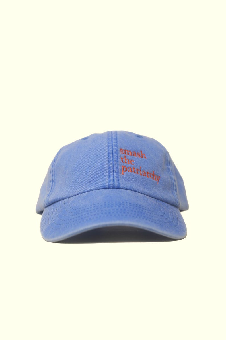 Op de afbeelding: Lichtblauwe baseballcap met de tekst "smash the patriarchy" in oranje geborduurd. De cap heeft een gebogen klep en een zes-panelen ontwerp, geschikt voor casual gebruik.