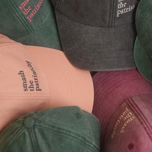 Op de afbeelding: Een verzameling baseballpetten in verschillende kleuren, waaronder donkergroen, poederroze en antracietgrijs. Elke pet heeft de geborduurde tekst "smash the patriarchy" in een contrasterende kleur. De petten zijn gemaakt van een zachte, getextureerde stof.