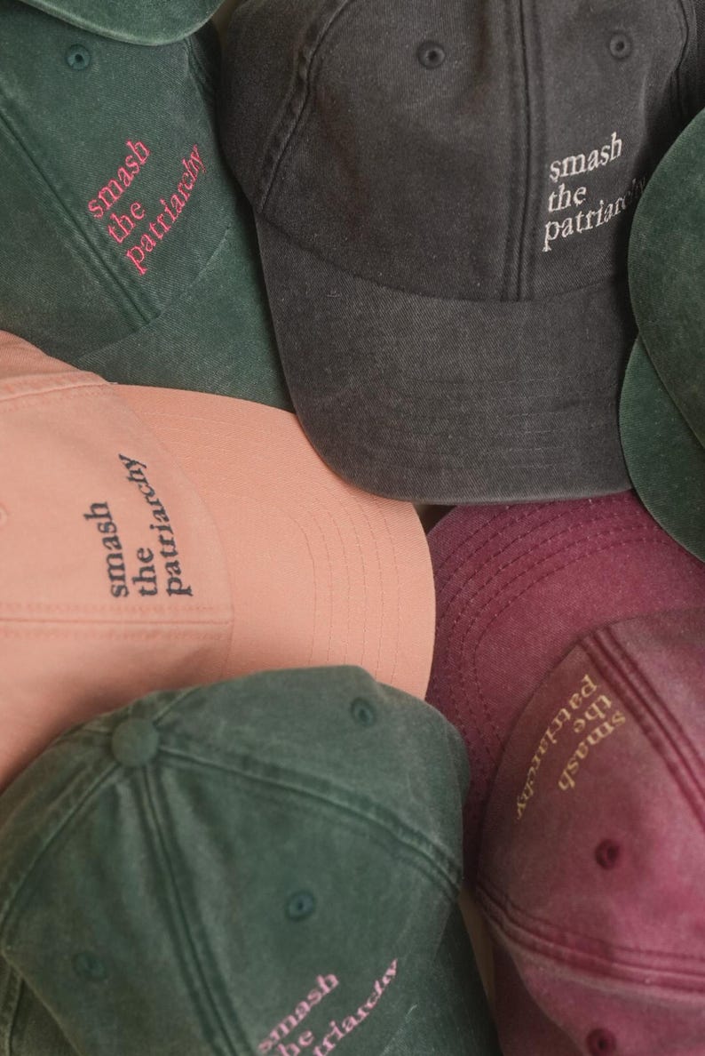 Op de afbeelding: Een verzameling baseballpetten in verschillende kleuren, waaronder donkergroen, poederroze en antracietgrijs. Elke pet heeft de geborduurde tekst "smash the patriarchy" in een contrasterende kleur. De petten zijn gemaakt van een zachte, getextureerde stof.