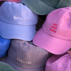 Puede incluir: Una colección de gorras de béisbol en varios colores, incluyendo rosa, azul y verde. Cada gorra presenta texto bordado como "baby", "bananas" y "not your baby". Las gorras están hechas de una tela suave y son adecuadas para uso casual.
