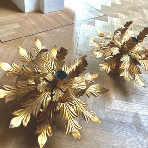 Puede incluir: Dos candelabros dorados ornamentados, cada uno con un diseño de hojas detallado. Los candelabros tienen un enchufe central para una vela y están colocados sobre un suelo de madera clara. El diseño evoca un arreglo floral.