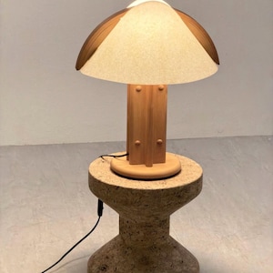 May include: A table lamp with a light-colored, textured, conical shade and a wooden base featuring a central column with four vertical fins. The lamp sits on a cylindrical, hourglass-shaped, speckled beige stone pedestal with a black power cord.