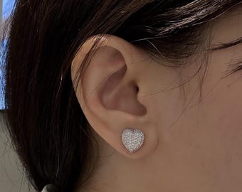 May include: A close-up of a heart-shaped stud earring. The earring is covered in small, sparkling stones, creating a shimmering effect. The earring is set against the backdrop of a person's ear and hair.