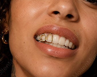 May include: A gold tooth grill set featuring two individual caps for adjacent teeth. One cap has a triangular frame with two small embedded stones, while the other is a teardrop-shaped frame accented with a row of five small clear stones along the base.
