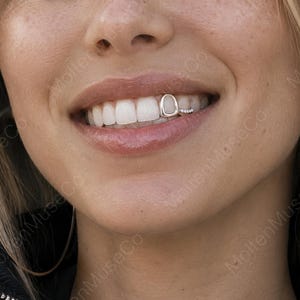 May include: A silver tooth grill consisting of a single smooth, polished cap on one tooth and a separate, smaller adjacent piece featuring a row of five tiny, clear, sparkling stones set into a thin, metallic band, worn on the upper teeth.