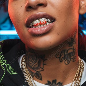 Peut inclure: Gros plan sur la bouche d'une personne avec des grillz incrustées de diamants avec un motif de flamme rouge. La personne a un tatouage de rose sur le cou et le mot "Chupito". Un collier en chaîne dorée est également visible.