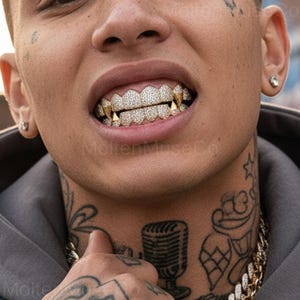 Könnte beinhalten: Nahaufnahme des Mundes einer Person mit diamantbesetzten Goldzähnen und Goldfangzähnen. Die Person hat Tätowierungen an Hals und Gesicht und trägt eine Goldkette. Die Zähne sind der Fokus des Bildes.