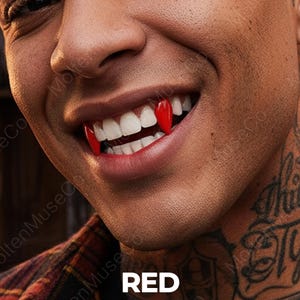 May include: Close-up of a person's smiling mouth with bright white teeth and two red vampire fangs. The word "RED" is displayed in white text at the bottom of the image. The person has tattoos on their neck.