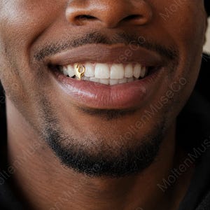 May include: A single gold tooth grill cap featuring a stylized, curved cutout design. The small, polished gold accessory is fitted onto an upper lateral incisor tooth, creating a distinct metallic accent against the natural white teeth.
