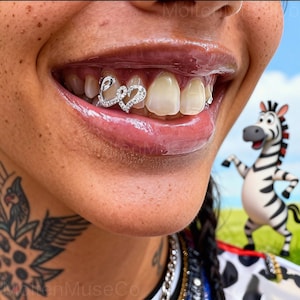 May include: A close-up of a person's smile, featuring silver dental grillz spelling "Love" with rhinestones. The lips are glossy, and a cartoon zebra is in the background.