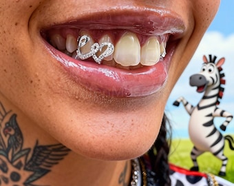 May include: Close-up of a person's smile, showcasing silver dental grillz spelling "Love" with embedded rhinestones. The lips are glossy, and a cartoon zebra is in the background.