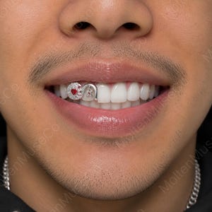 May include: A close-up of a person's smile, showcasing custom dental grills. The grill features a heart-shaped design with a red gem and a star, surrounded by clear stones, set in a silver metal.