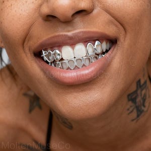 Può includere: Primo piano della bocca sorridente di una persona con grillz argentati. I grillz presentano forme di cuore e stella sui denti superiori e forme di cuore sui denti inferiori. I denti sono bianchi e le labbra sono di colore naturale.