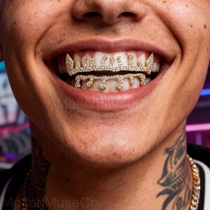 Può includere: Primo piano di una persona sorridente, che mostra grillz dorati. I grillz sono incastonati con strass scintillanti, con un design a goccia. Il testo "MoltenMuseCo" è visibile nell'angolo in basso a sinistra.
