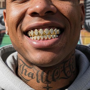 May include: Close-up of a person smiling, showcasing gold-colored teeth grills encrusted with small, sparkling diamonds. The grills are custom-fitted to the upper and lower teeth. The person has a tattoo on their neck and a diamond earring.