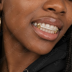 May include: A gold grillz piece worn on the upper teeth, featuring a horizontal bar set with a row of small, clear, sparkling stones. The design includes smooth, solid gold caps on the two outer teeth, creating a polished and metallic look.