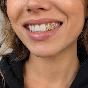 May include: A pair of gold tooth caps shaped like open hearts, worn on two adjacent upper teeth. The metallic gold covers the surface of the teeth, creating a distinct heart-shaped outline that frames the natural white enamel of the teeth.